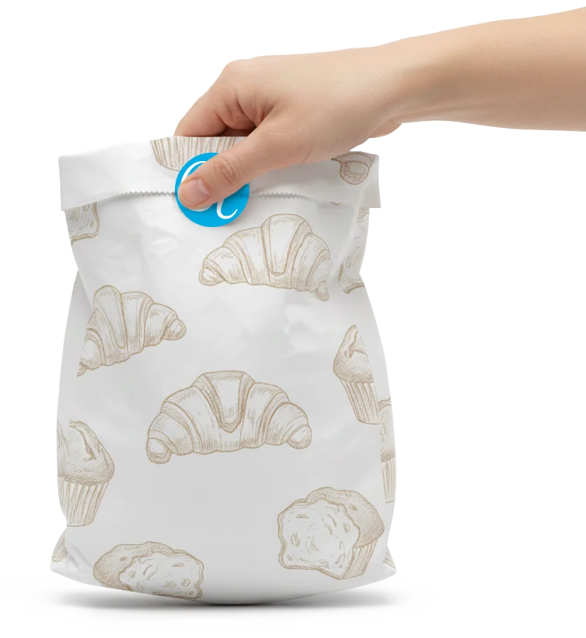 Sacchetto di carta decorata con disegni di croissant e muffin, sorretto da una mano e chiuso da un adesivo col logo di Luca Giavara